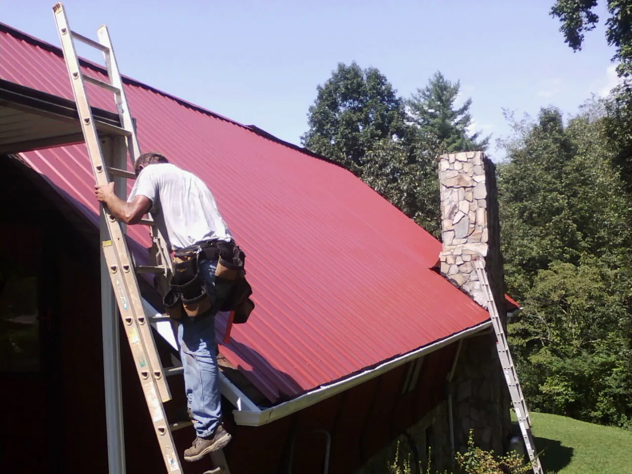Roofer on a ladder beside a red metal roof and stone chimney — Soffit & Fascia Repair by Roofing Brothers in Charleston